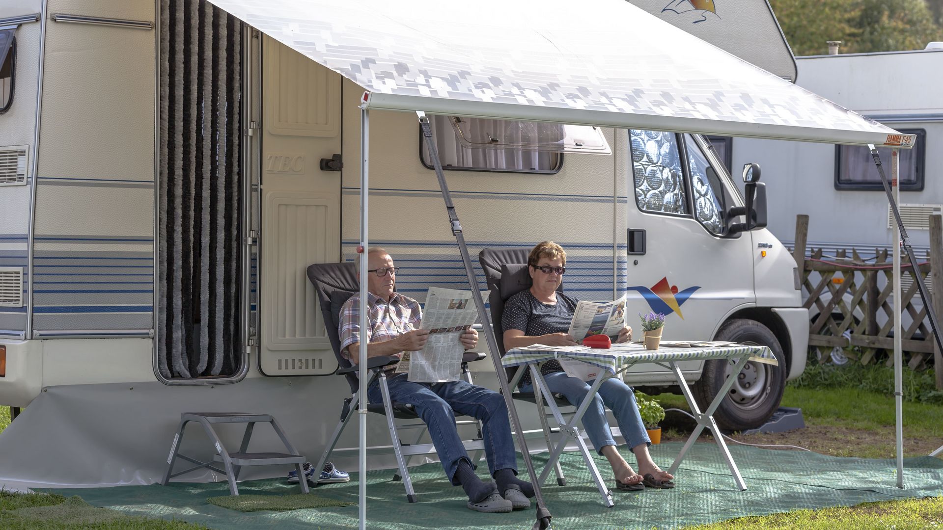 Camping & Stellplätze Das Bild zeigt ein Paar, sitzend auf Campingstühlen vor ihrem Wohnmobil. Beide lesen eine Zeitung.