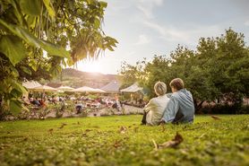 Paar beim Besuch des mosel musikfestivals © Rheinland-Pfalz Tourismus GmbH Man sieht eine Frau und einen Mann von hinten, die auf einer Wiese sitzen und auf einen Hof mit Menschen, Sonnenschirmen, stühlen und Tischen schauen.