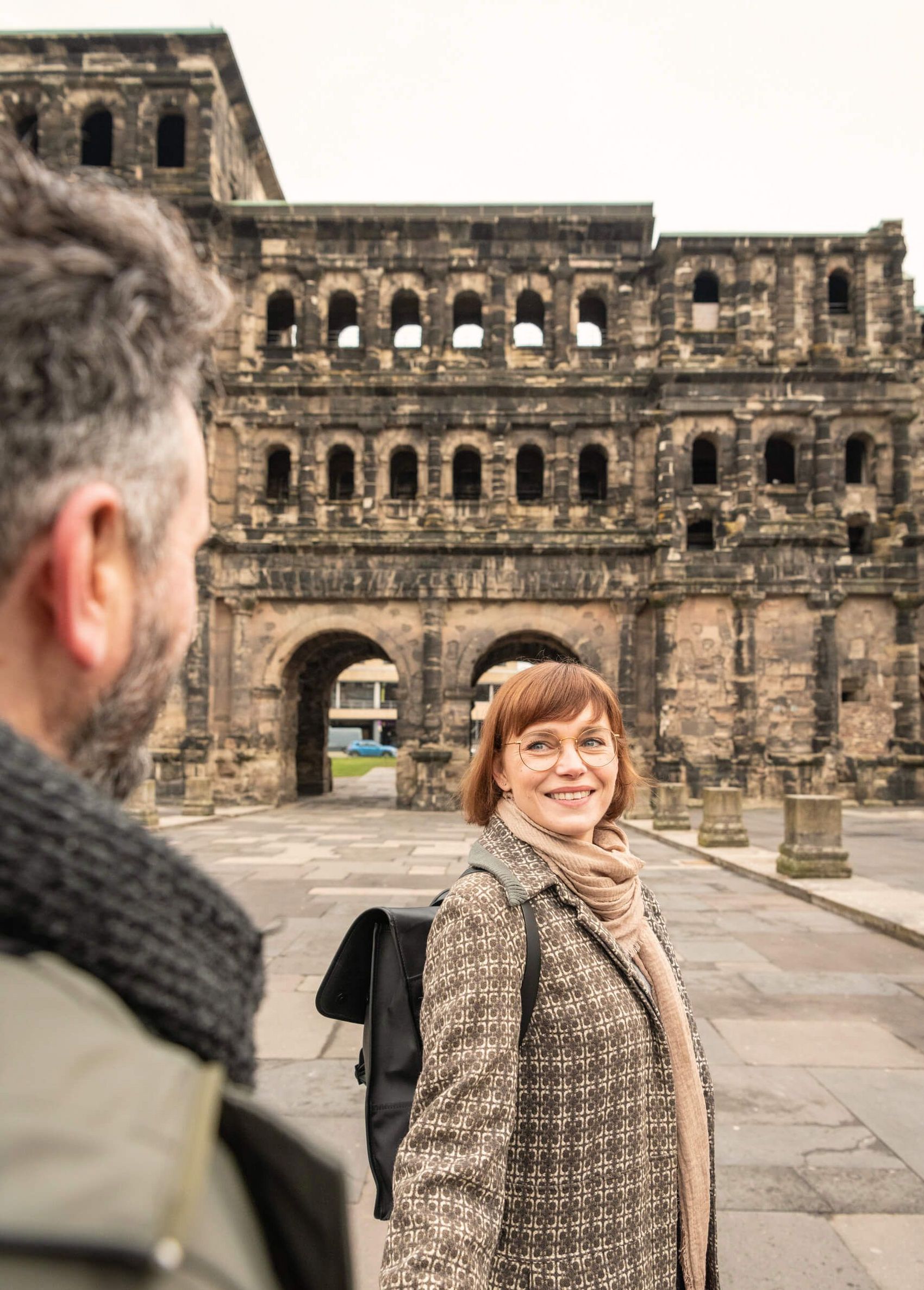Trier, Porta Nigra © D. Ketz / MLT GmbH Frau mit Mantel und schwarzem Rucksack vor Porta Nigra blickt über Schulter zu Mann