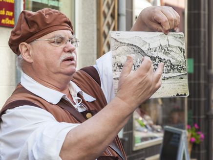 Riwigmännchen in Zell (Mosel) Auf dem Bild befindet sich ein älterer Herr in einem Kostüm. Er zeigt auf ein Bild, welches die Altstadt von Zell vor vielen Jahren darstellt. Er erklärt als Stadtgeist von Zell den Touristen vergangene Geschichten.
