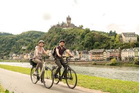 Radfahrer auf Radweg am Flussufer mit Burg im Hintergrund