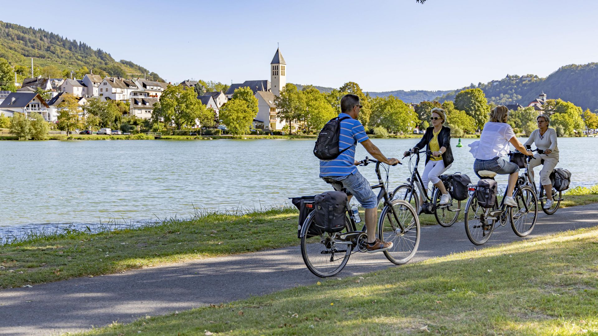 Mosel-Radweg Das Bild zeigt vier Personen auf ihren Fahrrädern, die sich auf einem Moselradweg entgegenkommen. Im Hintergrund ist der Moselort Bullay zu erkennen.