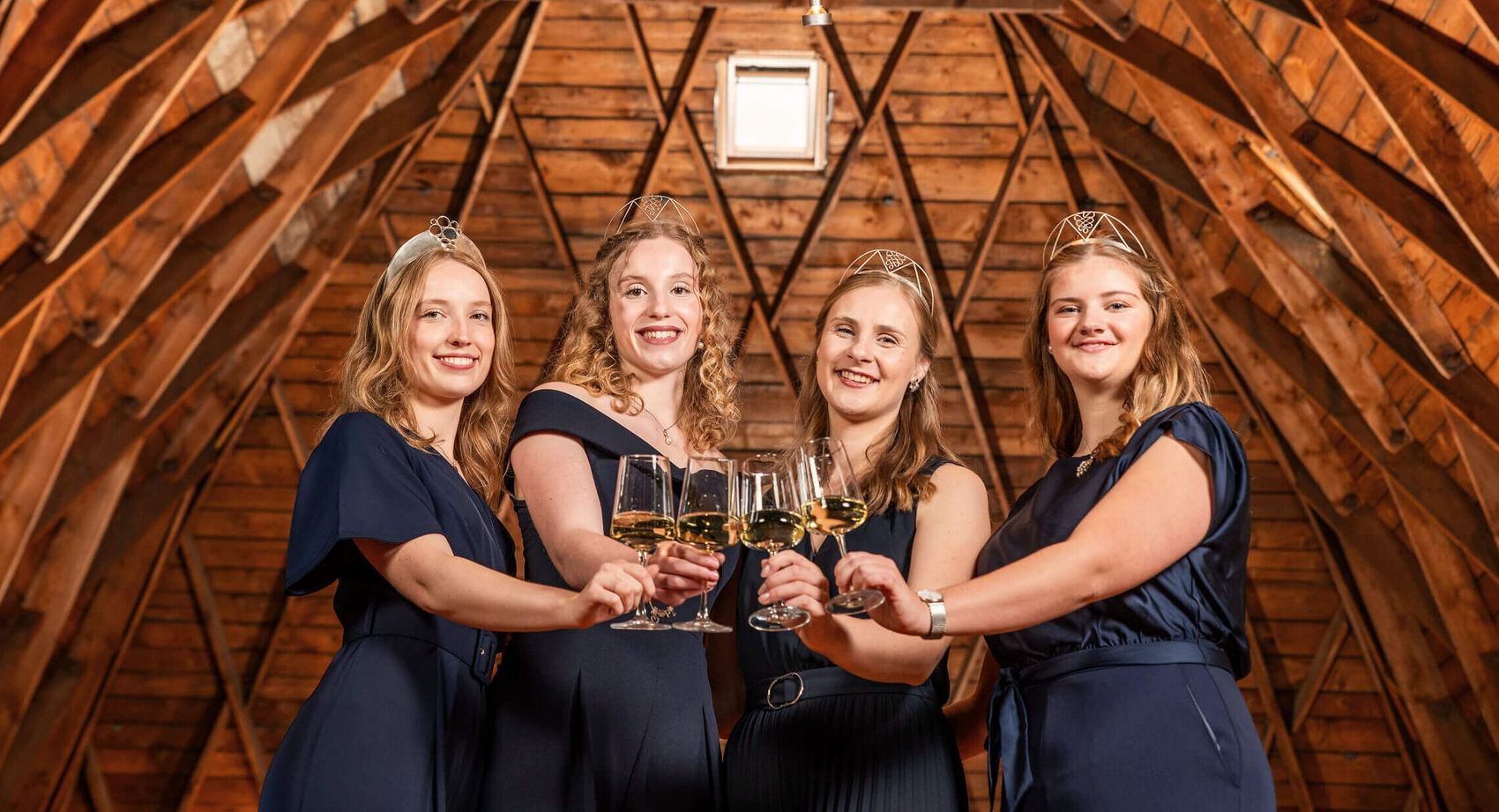 Moselweinhoheiten 2024/2025 © Chris Marmann/Moselwein e.V. Vier Frauen mit Kronen in blauen Kleidern schauen in die Kamera und stoßen mit Weingläsern an.