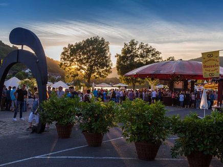 Weinfest Das Bild zeigt eine Aufnahme von dem Weinfest in Zell an der Mosel bei Sonnenuntergang. Im Vordergrund sind Pflanzen, sowie die Zeller Schwarze Katz als Torbogen zu erkennen und im Hintergrund befinden sich viele Menschen.